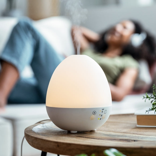 Shot of essential oil aroma diffuser humidifier diffusing water articles in the air while woman listening music lying on coach.