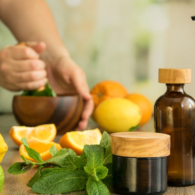 Tree glasses jars with essential oils against background of human hands pounding with pestle in wooden mortar and slices of orange, lemon, leaves of mint