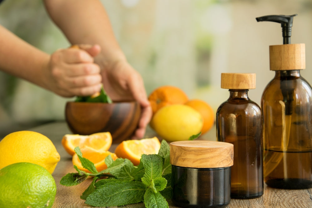 Tree glasses jars with essential oils against background of human hands pounding with pestle in wooden mortar and slices of orange, lemon, leaves of mint