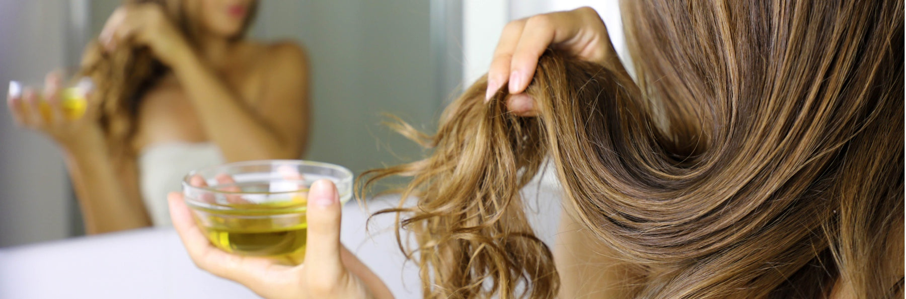 woman applying tamanu oil to hair in front of bathroom mirror in towel