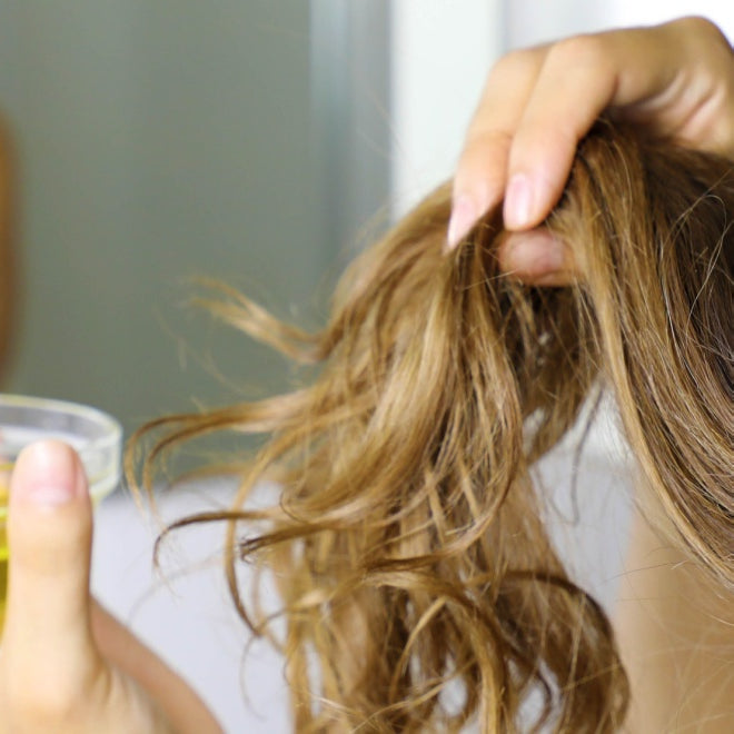 woman applying tamanu oil to hair in front of bathroom mirror in towel