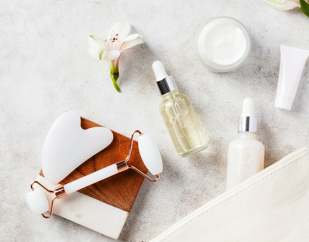Skincare products including a roller, bottles, and jars on a light surface with flowers.